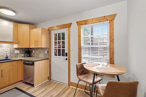 A compact kitchen with wooden cabinets, a built-in dishwasher, a small window with blinds, and a round table with two chairs. - Cozy King Condo Walk to Bojangles & Ovens (Charlotte)