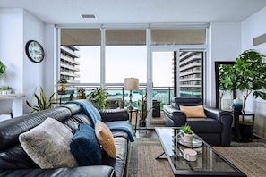 A modern living room with a black leather sofa, a glass coffee table, and a view of a cityscape through large windows.