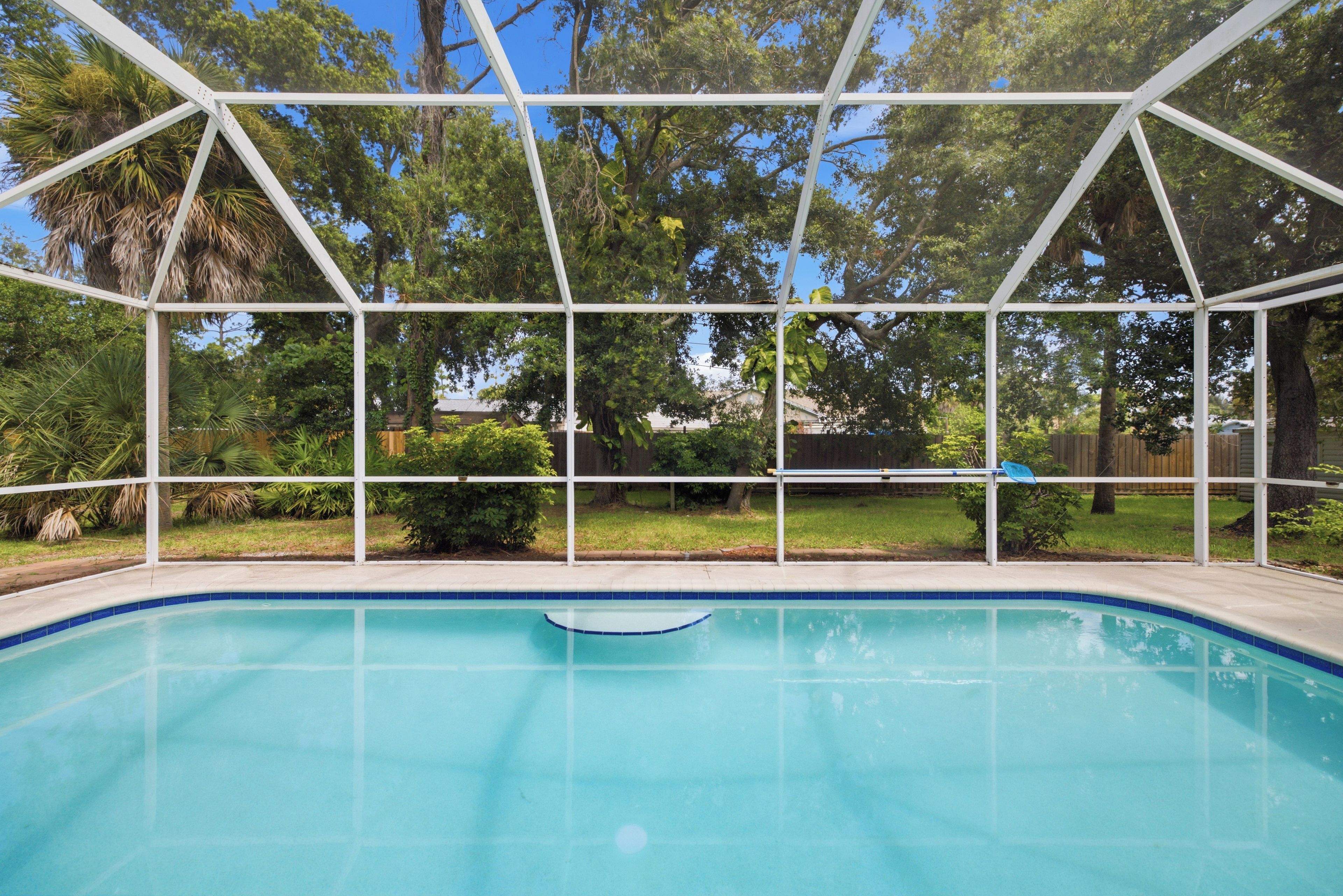 A clear blue swimming pool with a white safety fence, surrounded by lush greenery and trees.