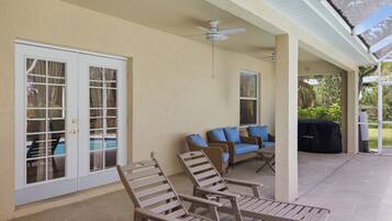 A covered patio area with two lounge chairs, a ceiling fan, and a sliding glass door leading to a pool.