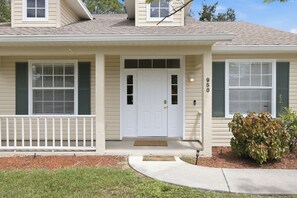 A two-story house with a front porch, a white door, and a small front yard with a shrub.