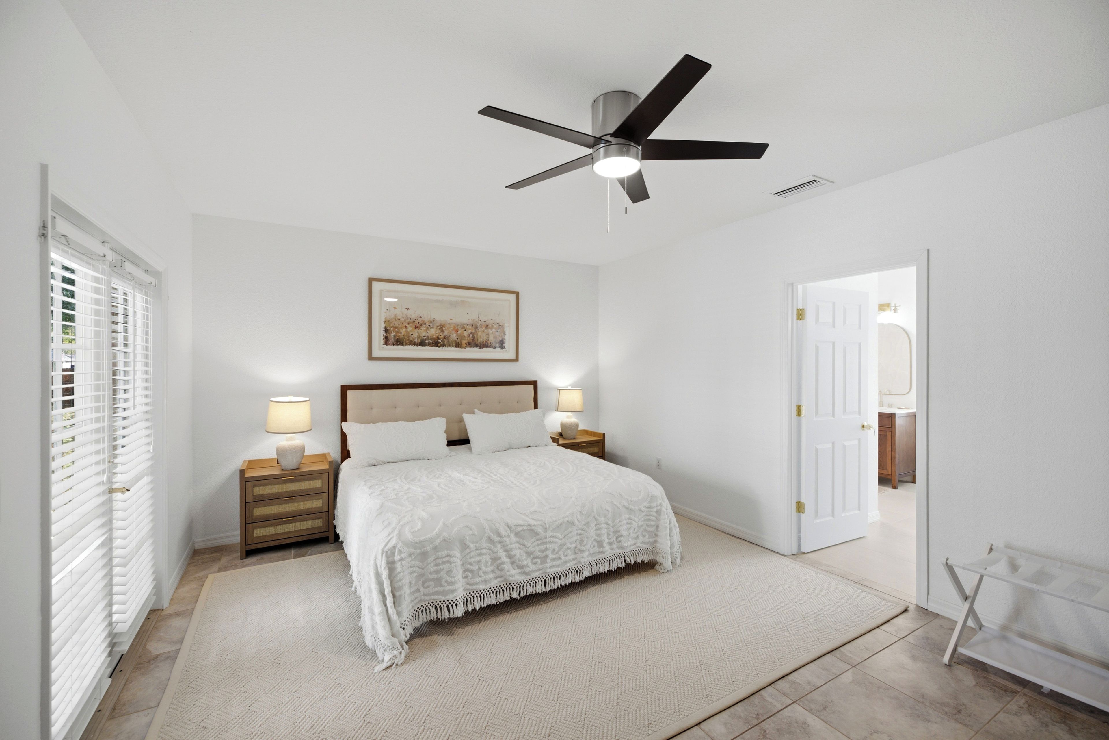 A bedroom with a bed, two bedside tables with lamps, a ceiling fan, a door, and a window with blinds.