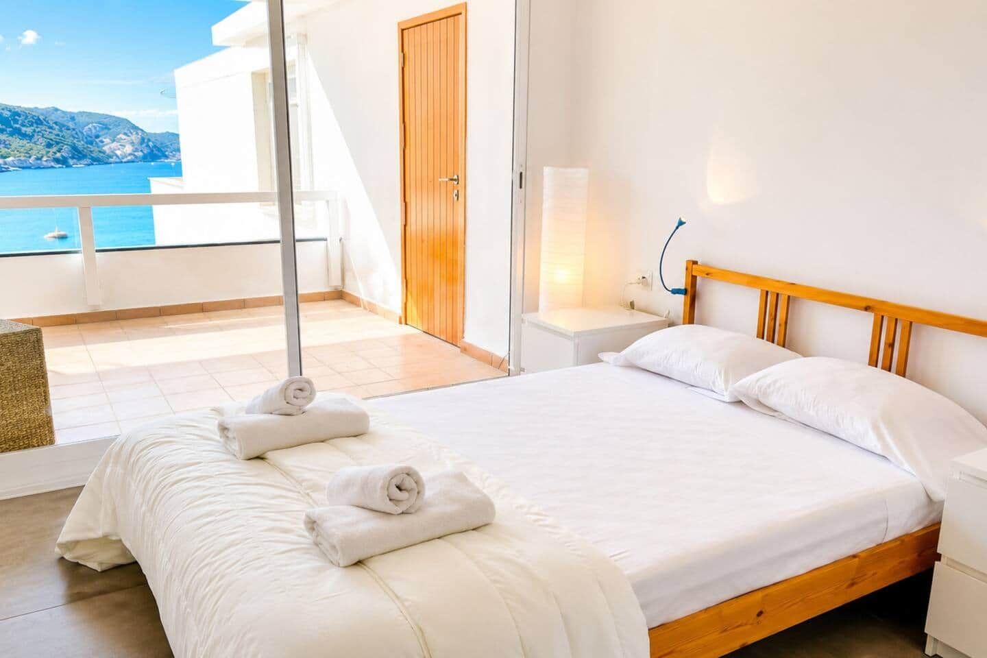 Habitación de hotel con cama, cabecera de madera, mesita de noche, lámpara, vista al mar y balcón.