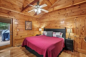 A wooden cabin bedroom with a bed, a nightstand, a lamp, a ceiling fan, and a window with a view of the outdoors. - Private Theater & Hot tub with Arcade and 3 King Beds (Sevierville)