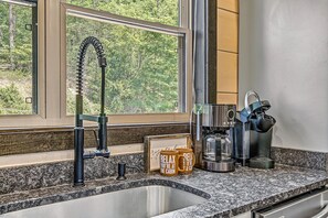 A kitchen with a granite countertop, a stainless steel sink, a coffee maker, and a window with a view of trees. - Private Indoor Pool + Theater + Game Room + Fire Pit | Sleeps 12 (Sevierviille)