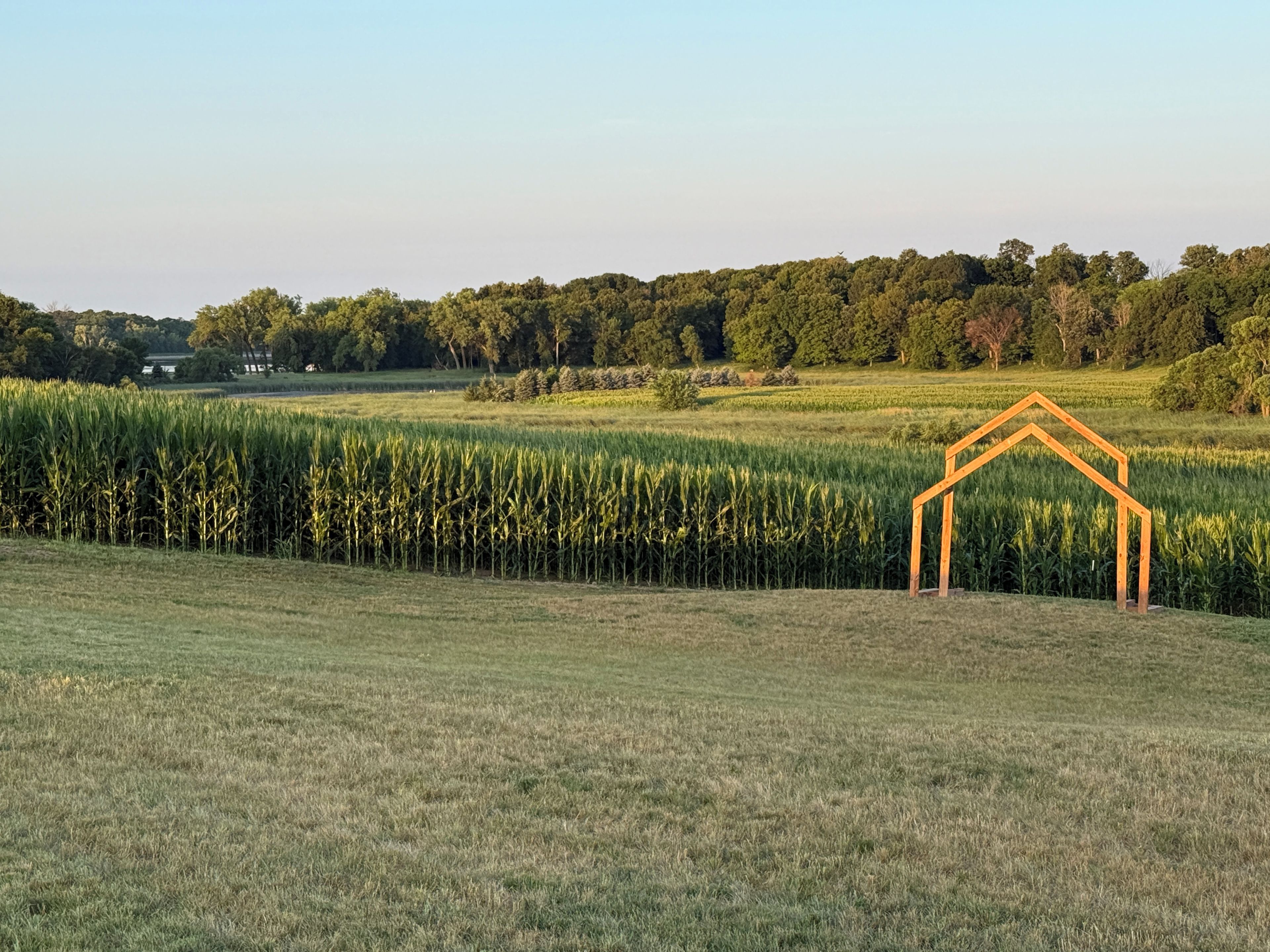Ländliche Landschaft mit einer hölzernen Struktur, die einem Haus ähnelt, im Vordergrund, umgeben von Feldern und Bäumen im Hintergrund.