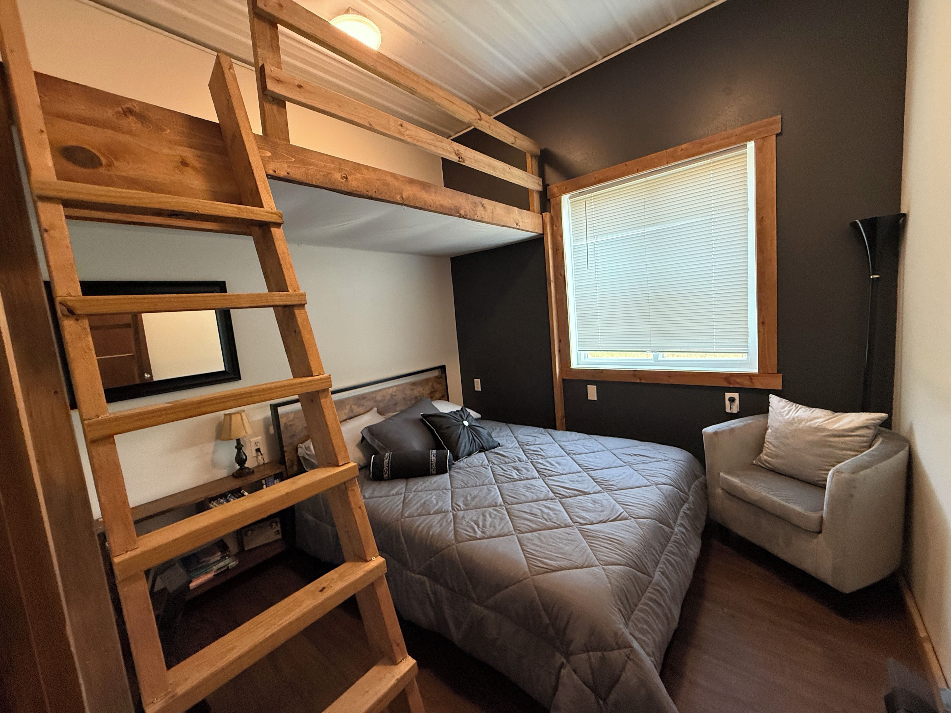 A bedroom with a bunk bed, a ladder, a window with blinds, a bedside table, and a chair.