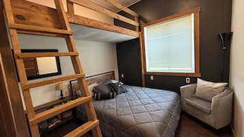 A bedroom with a bunk bed, a ladder, a window with blinds, a bedside table, and a chair.