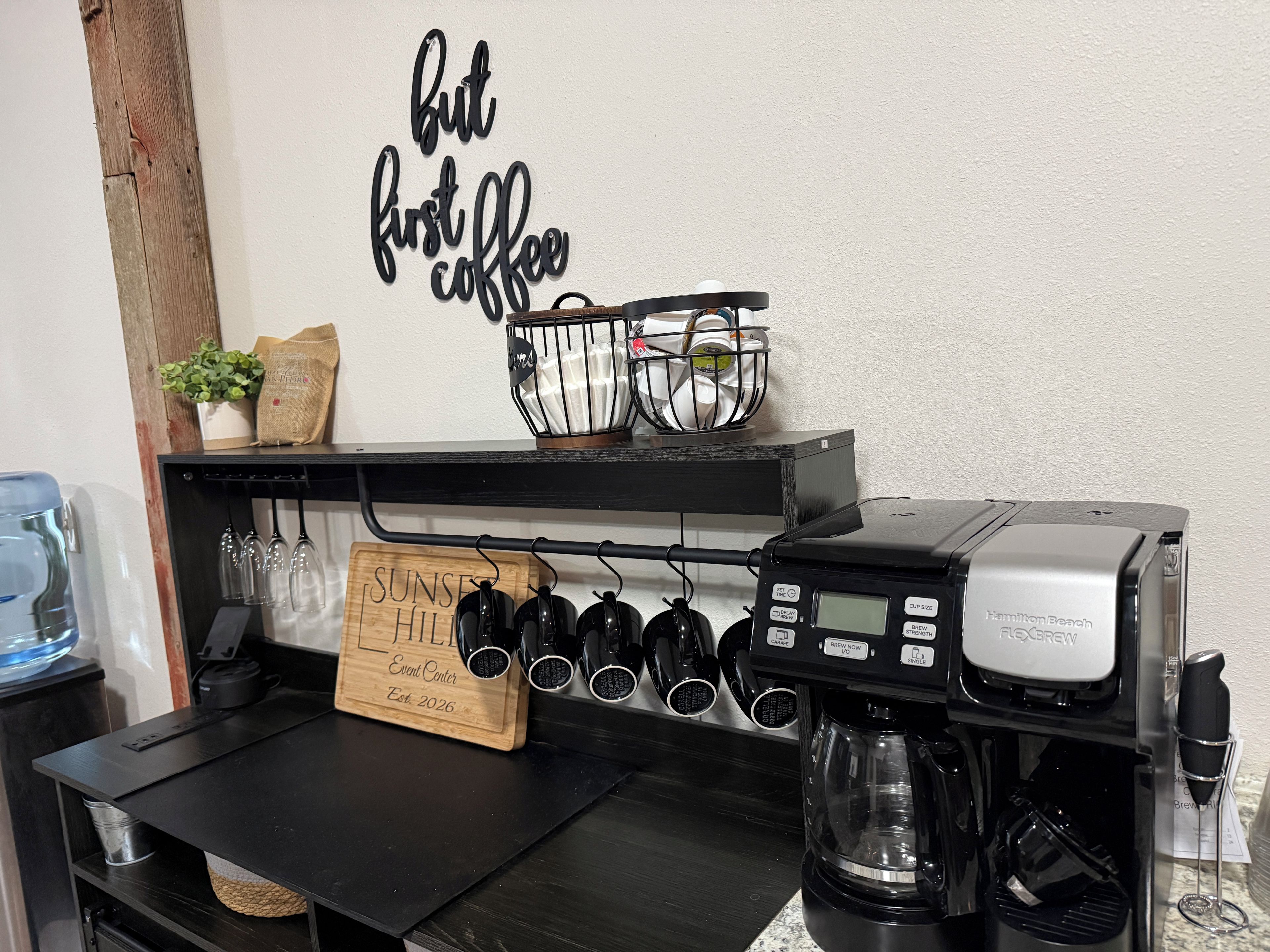 A coffee station with a Hamilton Beach coffee maker, a sign reading 'Sunset Hill Event Center Est. 2026,' and a wall sign that says 'but first coffee.'