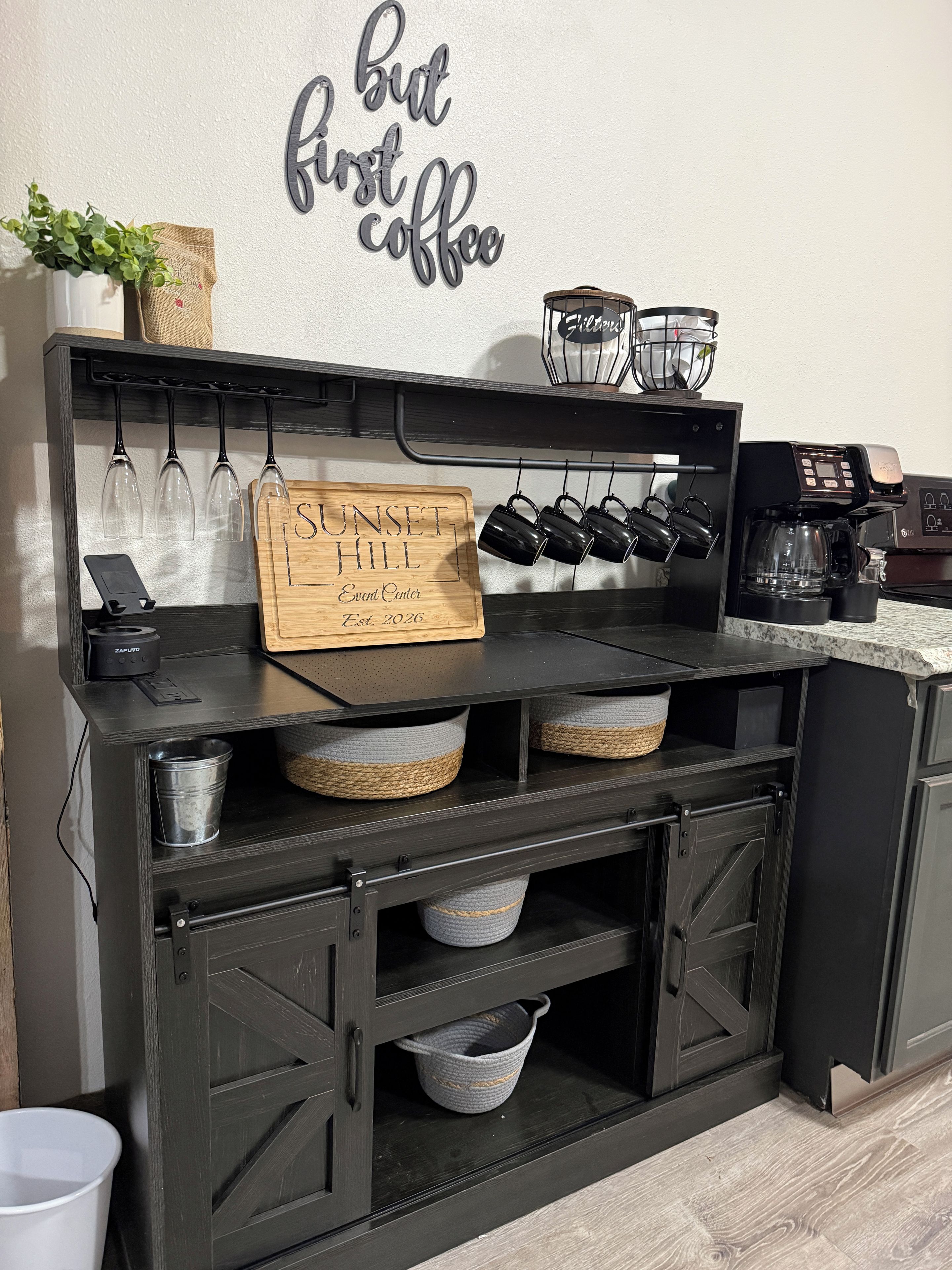 A kitchen counter with a coffee machine, wine glasses, and a sign that reads 'SUNSET HILL Event Center.'