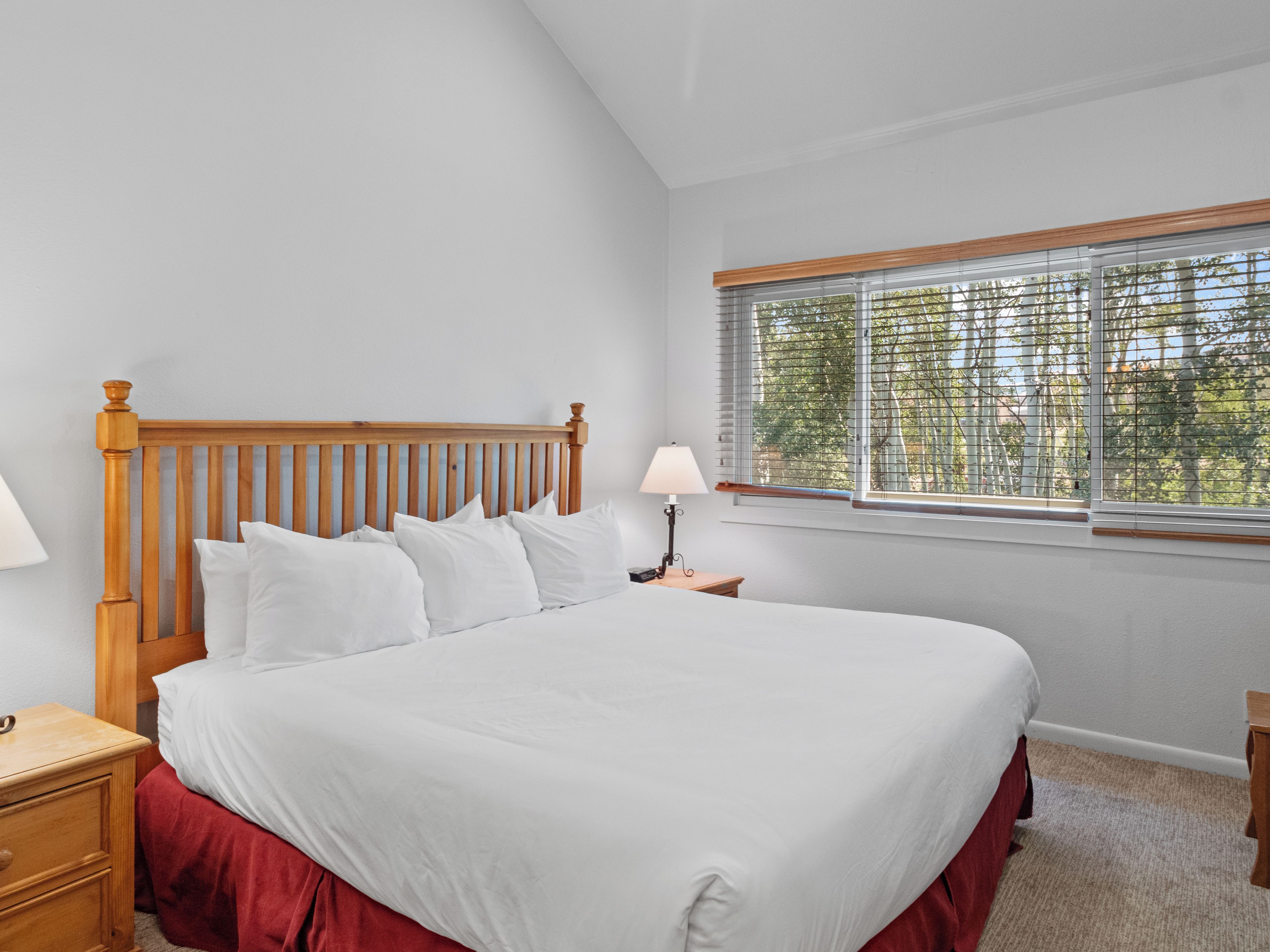 A bedroom with a wooden headboard, a large bed with white bedding, a nightstand with a lamp, and a window with blinds.