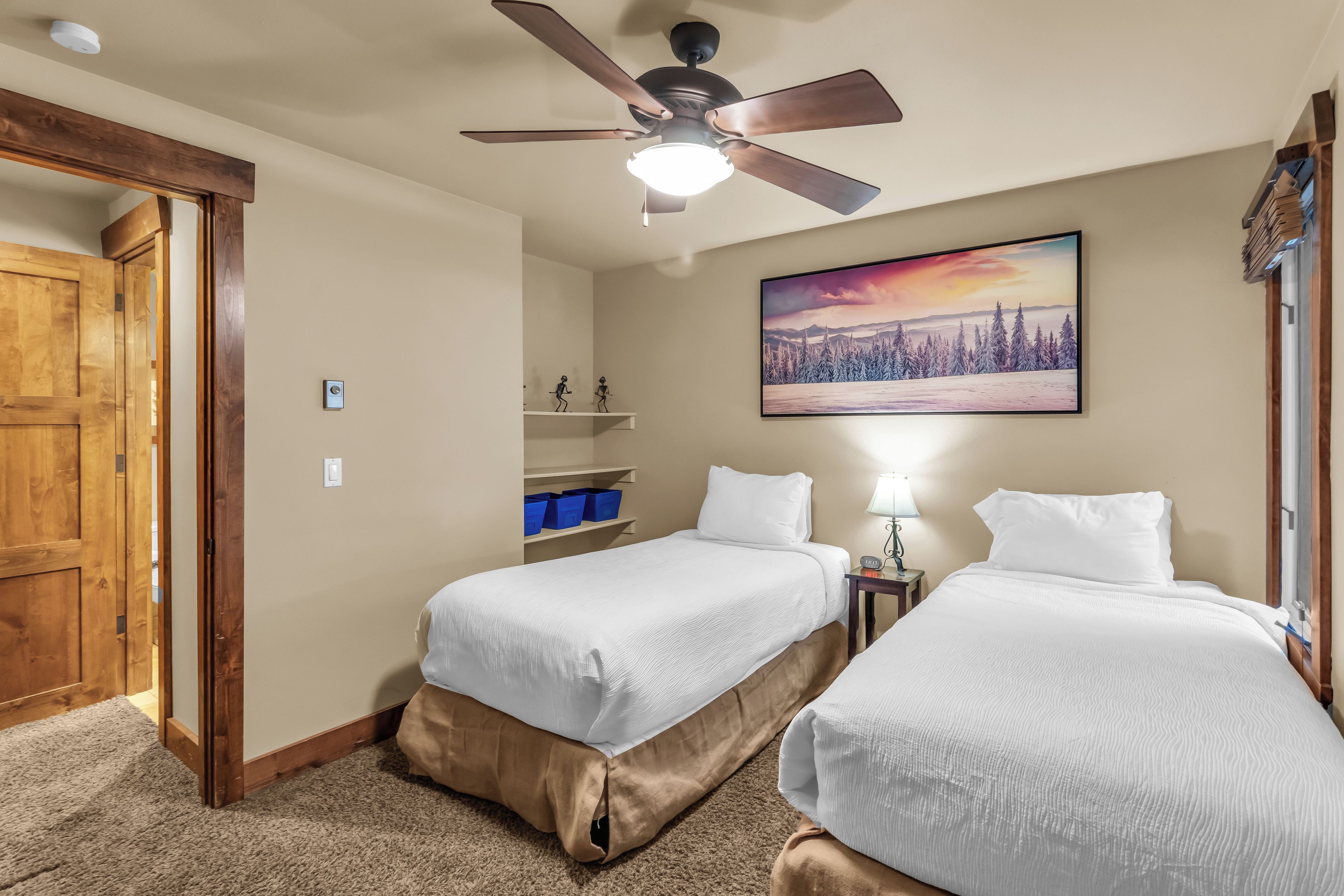 A hotel room with two beds, a ceiling fan, a painting of a winter landscape, and a wooden door.