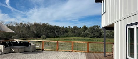Een houten terras met buitenmeubilair, een wit gebouw met grote ramen en uitzicht op een bebost gebied.