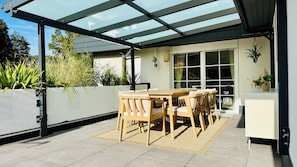 An outdoor patio with a glass roof, a wooden table and chairs, and potted plants.