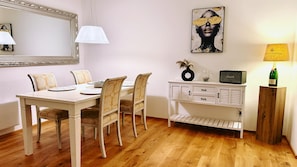 Un coin repas avec une table blanche, quatre chaises, une lampe, une œuvre d’art encadrée, un vase décoratif, une radio et une bouteille de champagne.