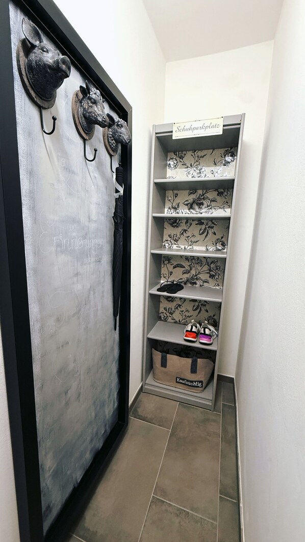 Un couloir étroit avec une porte ornée de têtes d’animaux en métal décoratives et de crochets. À côté se trouve un meuble de rangement pour chaussures, doté d’étagères à motifs floraux et d’un petit marchepied portant l’étiquette « Kaufpflichtig ».