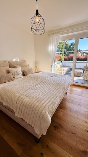 A bedroom with a large bed, a geometric pendant light, and a view of outdoor wicker furniture through a glass door.