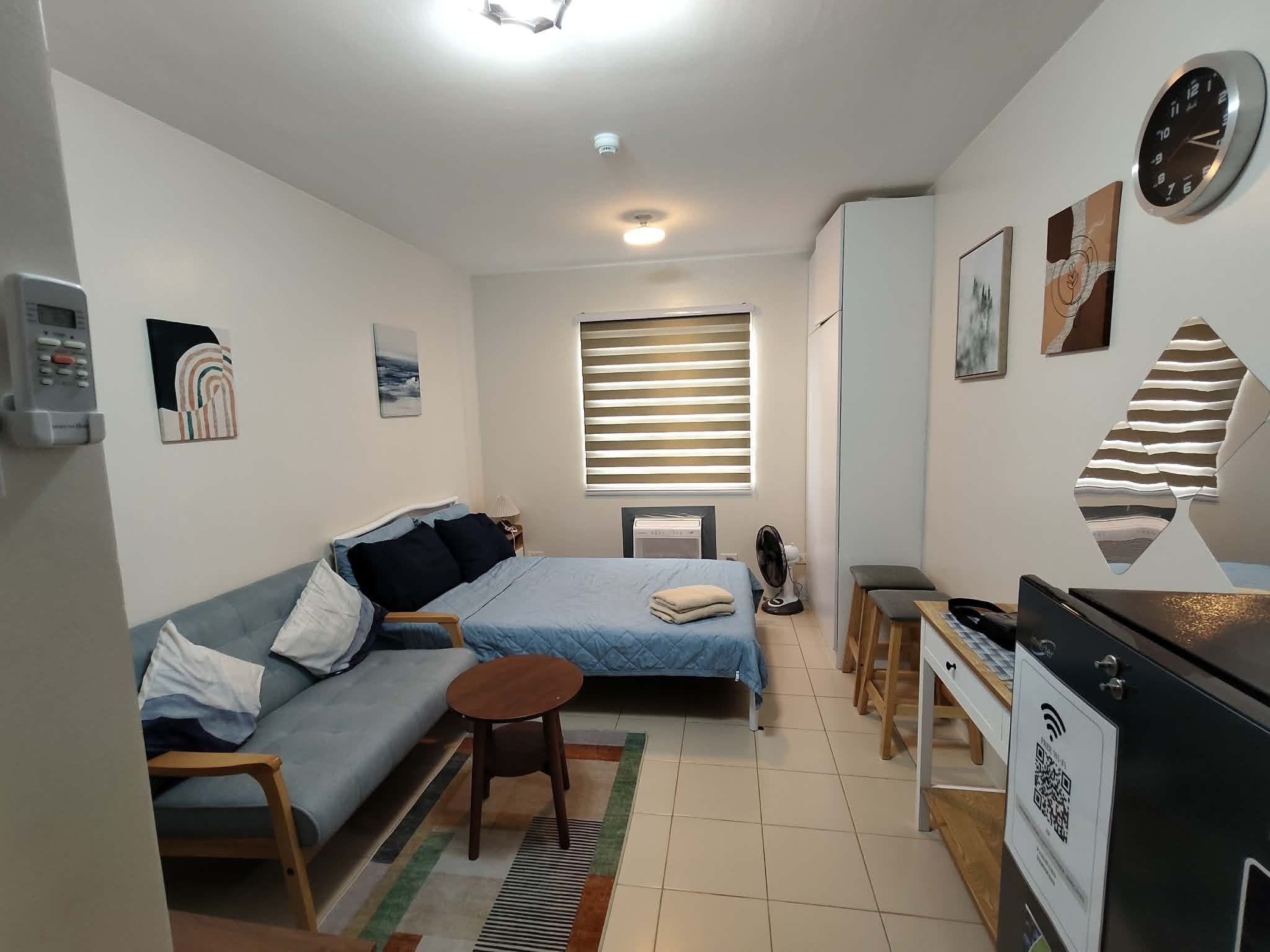 Un espacio habitable compacto con sofá, cama, una mesita, escritorio, radiador y un reloj en la pared.