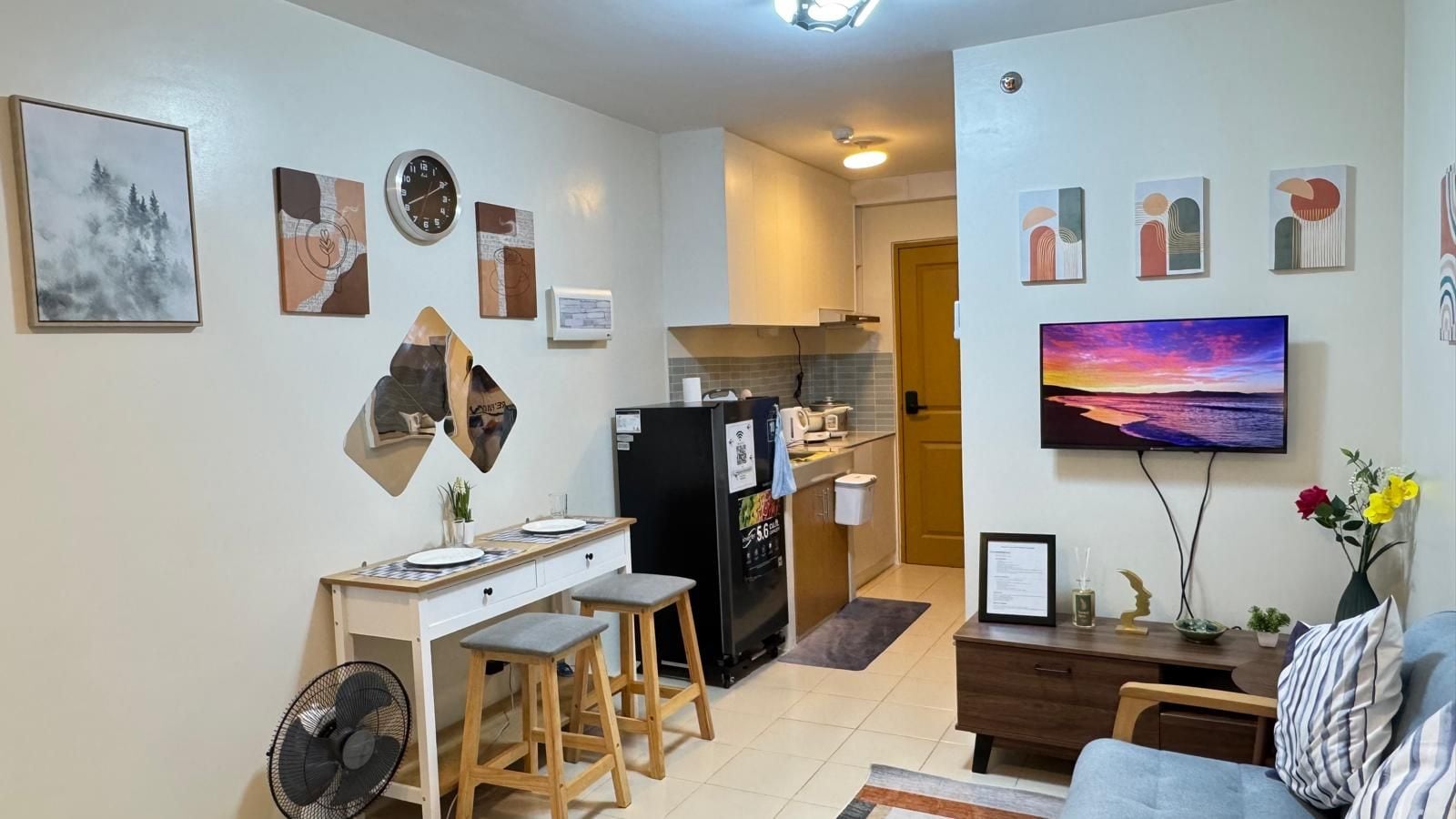 Ein Wohnzimmer mit einer Couch, einem Couchtisch und einem Fernseher. Es gibt eine Küchenzeile mit einem Kühlschrank, einer Mikrowelle und Schränken.