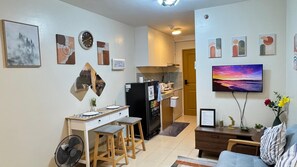 Ein Wohnzimmer mit einer Couch, einem Couchtisch und einem Fernseher. Es gibt eine Küchenzeile mit einem Kühlschrank, einer Mikrowelle und Schränken.