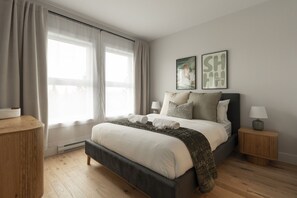 Une chambre à coucher comprenant un lit, deux tables de chevet, une fenêtre avec des rideaux et deux tableaux accrochés au mur.