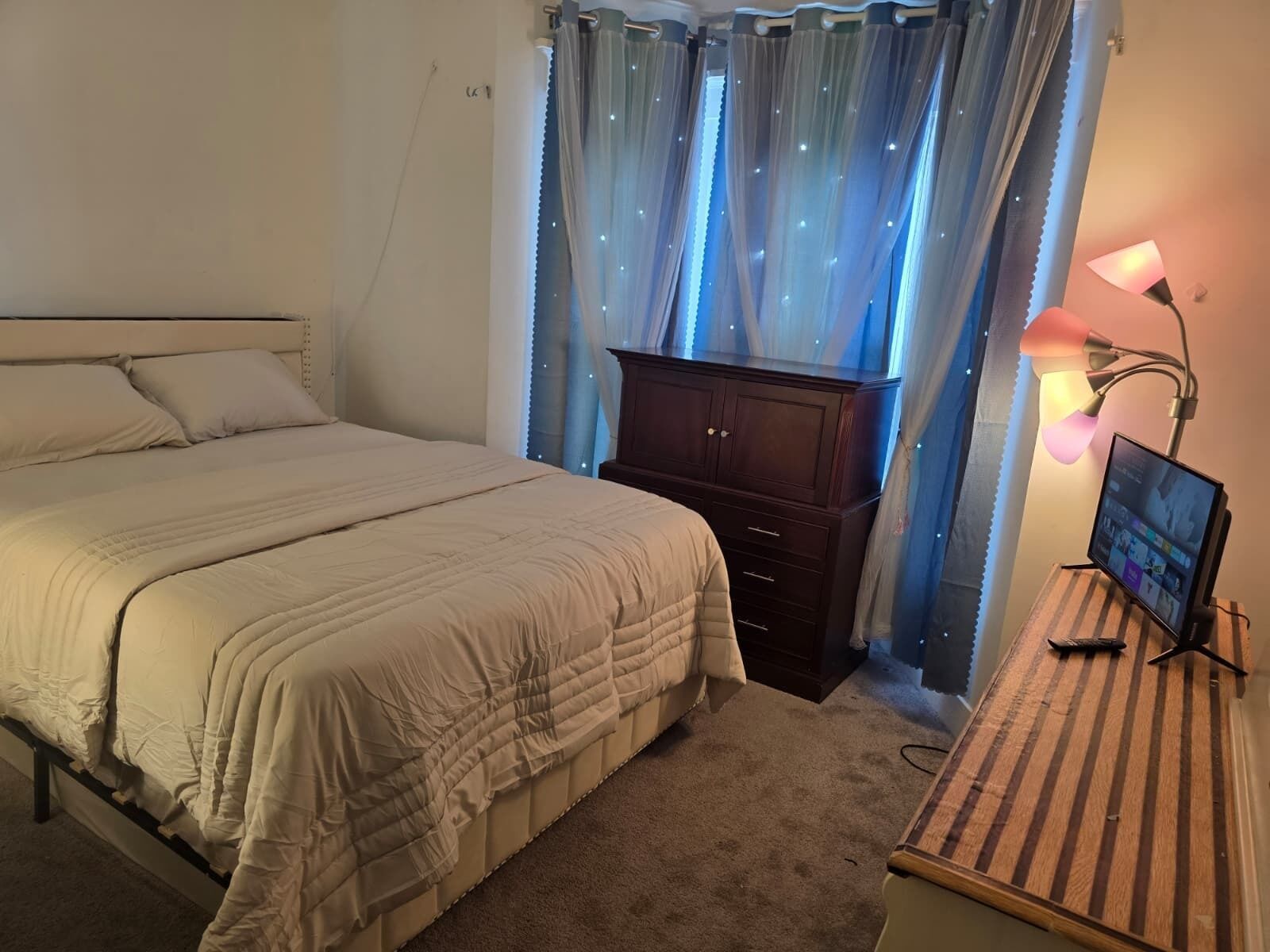 A bedroom with a bed, a dresser, a desk with a computer, and a window with curtains.
