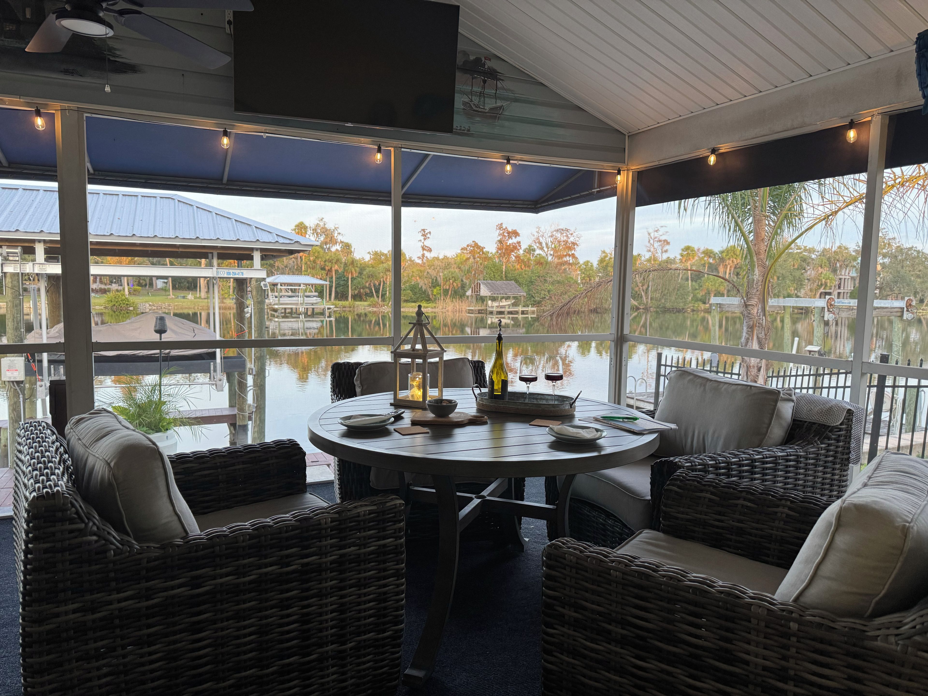 A covered outdoor seating area with wicker furniture, a table with a bottle and glasses, and a view of a water body and trees.