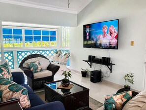 Ein Wohnzimmer mit an der Wand montiertem Fernseher, einer Couch, einem Couchtisch und einem Fenster mit Blick auf Grünflächen.