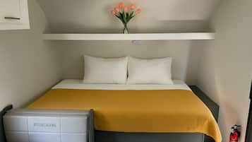 A compact hotel room with a bed, a bedside table, a chair, a vase with flowers, and a fire extinguisher.