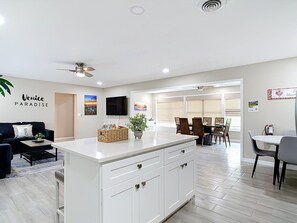 Eine moderne Küche mit einer weißen Kochinsel, ein Essbereich mit Tisch und Stühlen sowie ein Wohnzimmer mit Sofa und Fernseher.