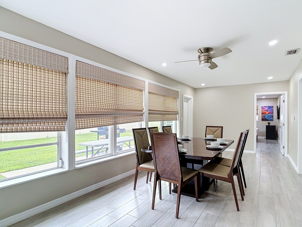A dining area with a table and chairs, large windows with blinds, a ceiling fan, and a hallway leading to another room.