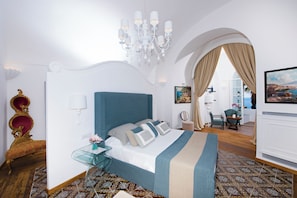 A hotel room with a large bed, a chandelier, a view through an arched doorway, and a dining area with a table and chairs. - Villa Evia by My Villa Collection (Positano)