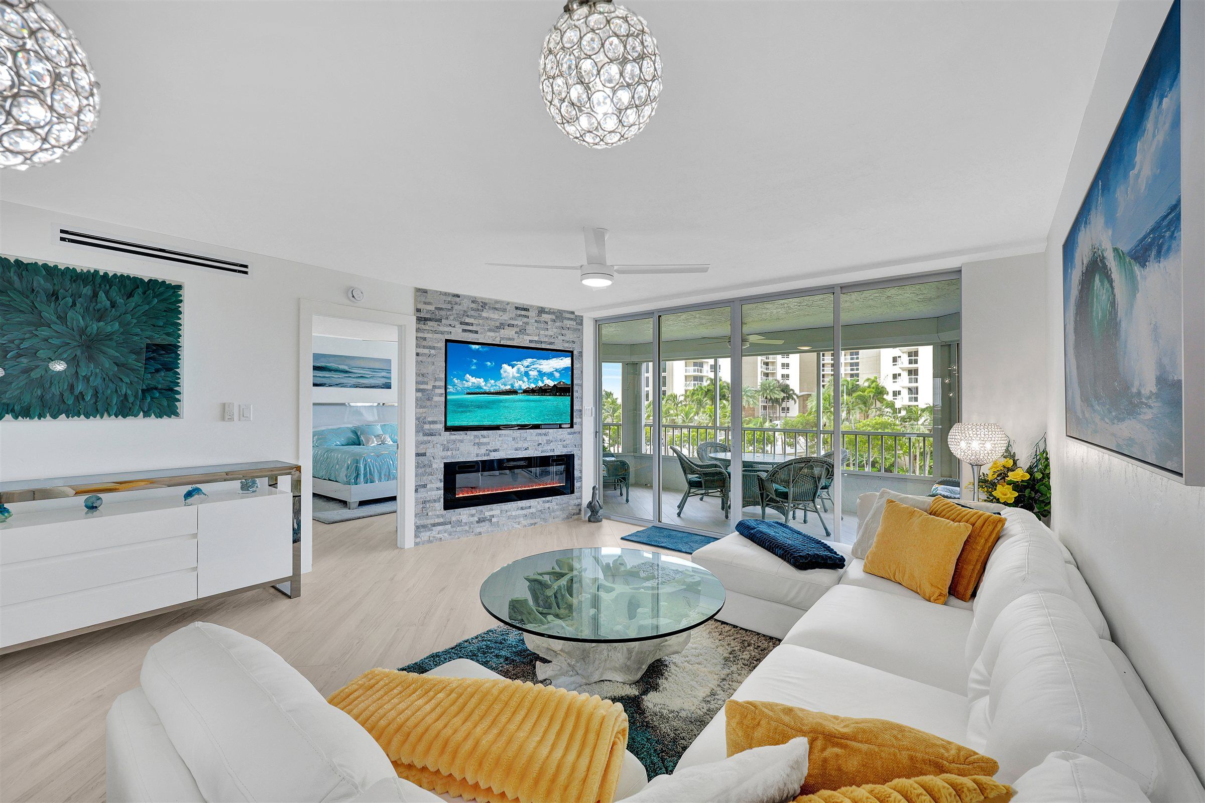 A modern living room with a flat-screen TV, a fireplace, a glass coffee table, and a balcony with outdoor furniture.