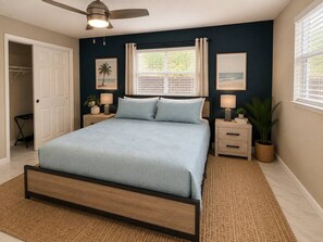 Un dormitorio con cama, mesitas de noche, ventilador de techo, ventana con cortinas y puerta.
