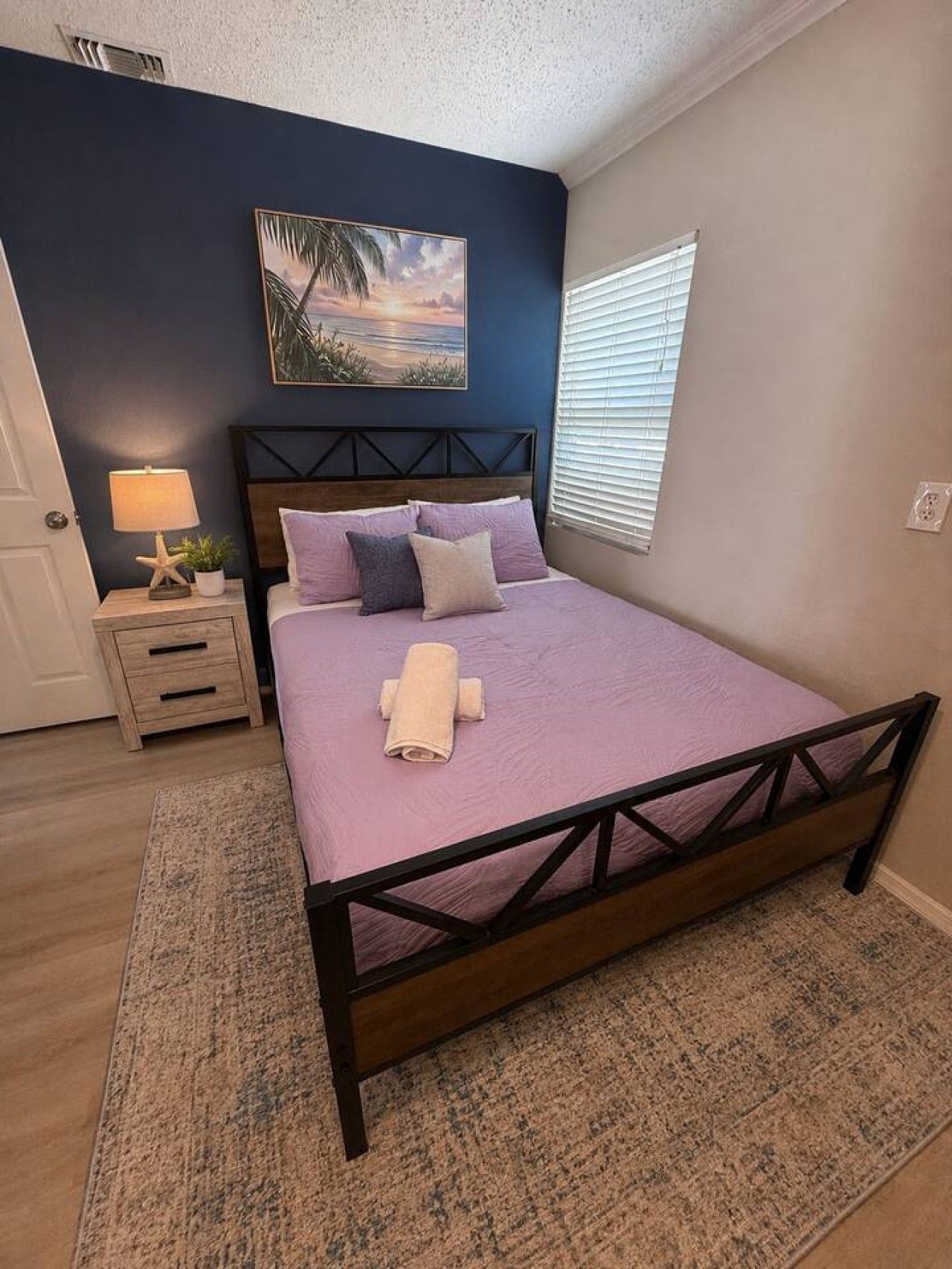 Une chambre à coucher avec un lit, une table de chevet, une lampe, un tableau et une fenêtre.