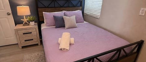 Quarto com cama, mesa de cabeceira, luminária, pintura e janela.