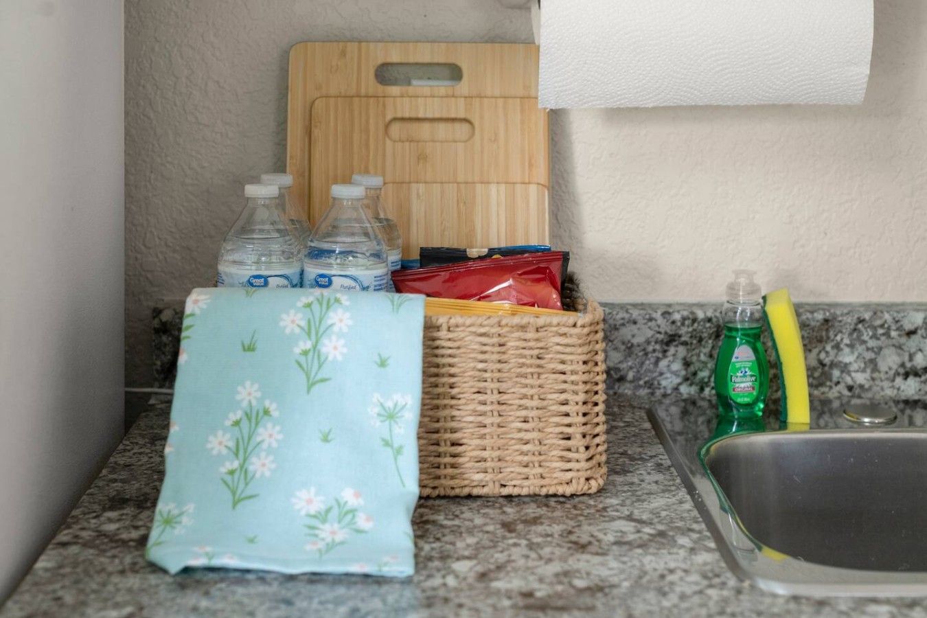 Una encimera de cocina con una tabla de cortar, botellas, una cesta, una botella de lavavajillas y una esponja.