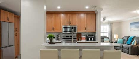 A modern kitchen with wooden cabinets, stainless steel appliances, a central island with bar stools, and a living area with a couch and lamp.