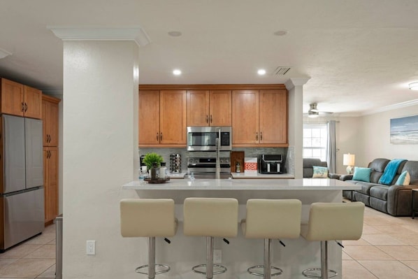 A modern kitchen with wooden cabinets, stainless steel appliances, a central island with bar stools, and a living area with a couch and lamp.
