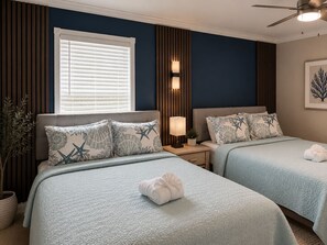 Quarto de hotel com duas camas, janela com persianas, ventilador de teto e luminária na parede.