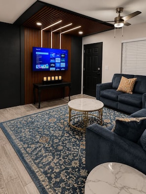 A modern living room with a flat-screen TV mounted on a wooden wall, a dark blue sofa, a round coffee table, and a patterned rug. - AMI Coastal Retreat Heated Pool 4BR 2BA Sleep 12 (Bradenton)