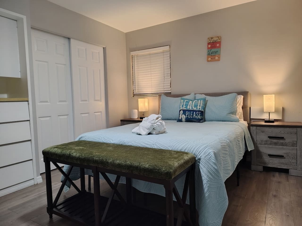 A bedroom with a bed, a bench, a nightstand, a lamp, and a door.