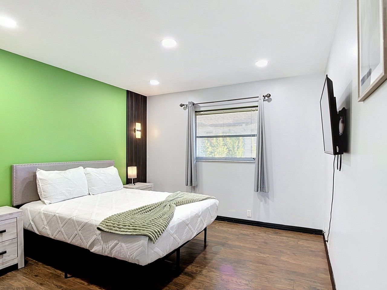 Une chambre moderne avec un lit, des tables de chevet, une fenêtre avec des stores et un téléviseur fixé au mur.