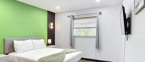 A modern bedroom with a bed, bedside tables, a window with blinds, and a wall-mounted TV.