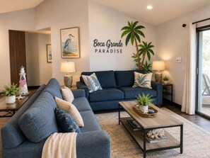 A living room with a sofa, coffee table, and decorative pillows. There is a wall with a palm tree and the text 'Boca Grande Paradise'.