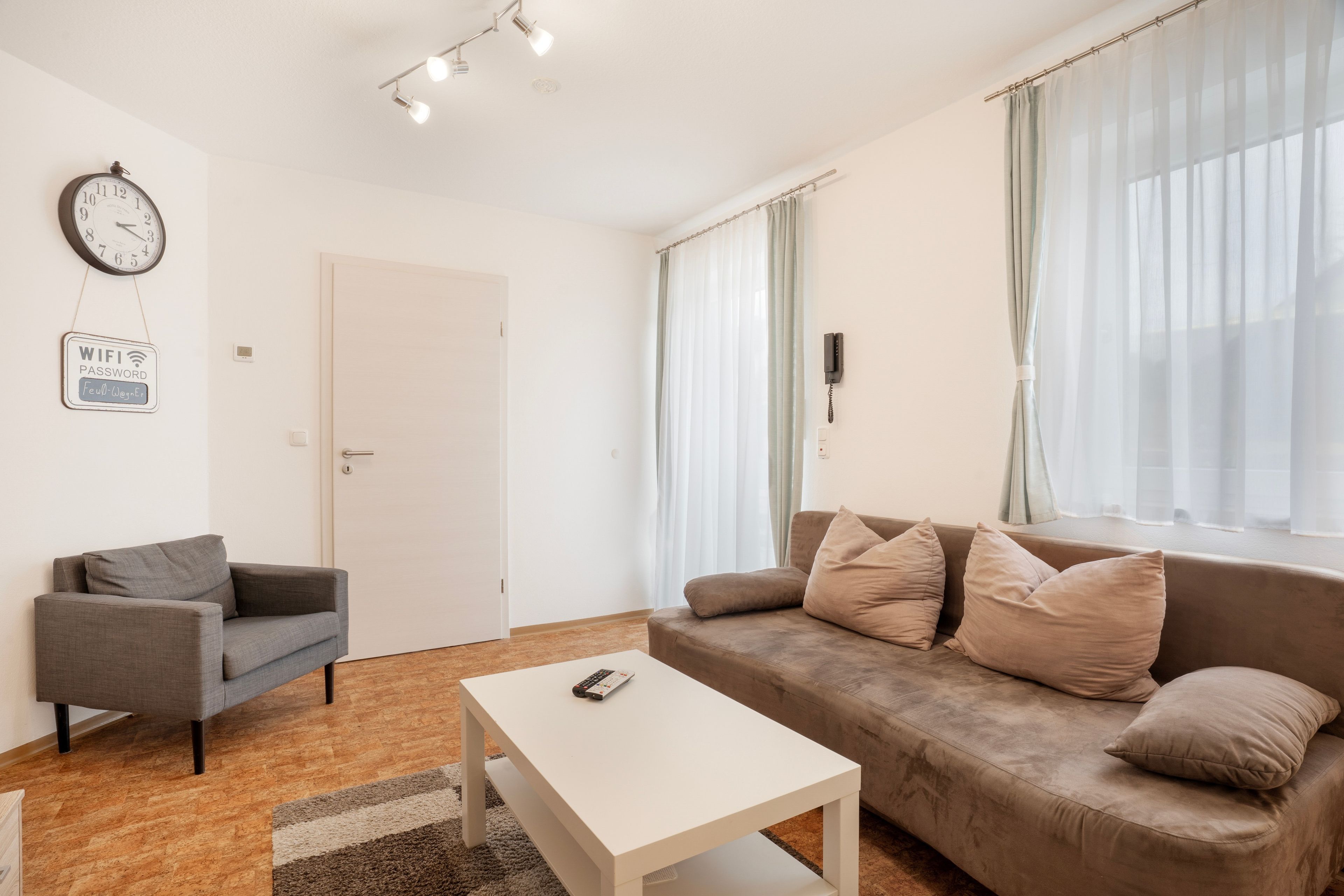 Een moderne woonkamer met een bank, een salontafel, een grijze fauteuil, een wandklok en een bordje met het WiFi-wachtwoord.