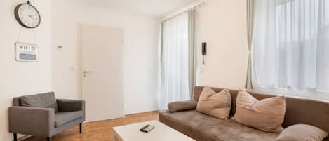 Un salon moderne avec un canapé, une table basse, un fauteuil gris, une horloge murale et une affiche indiquant le mot de passe du réseau Wi-Fi.