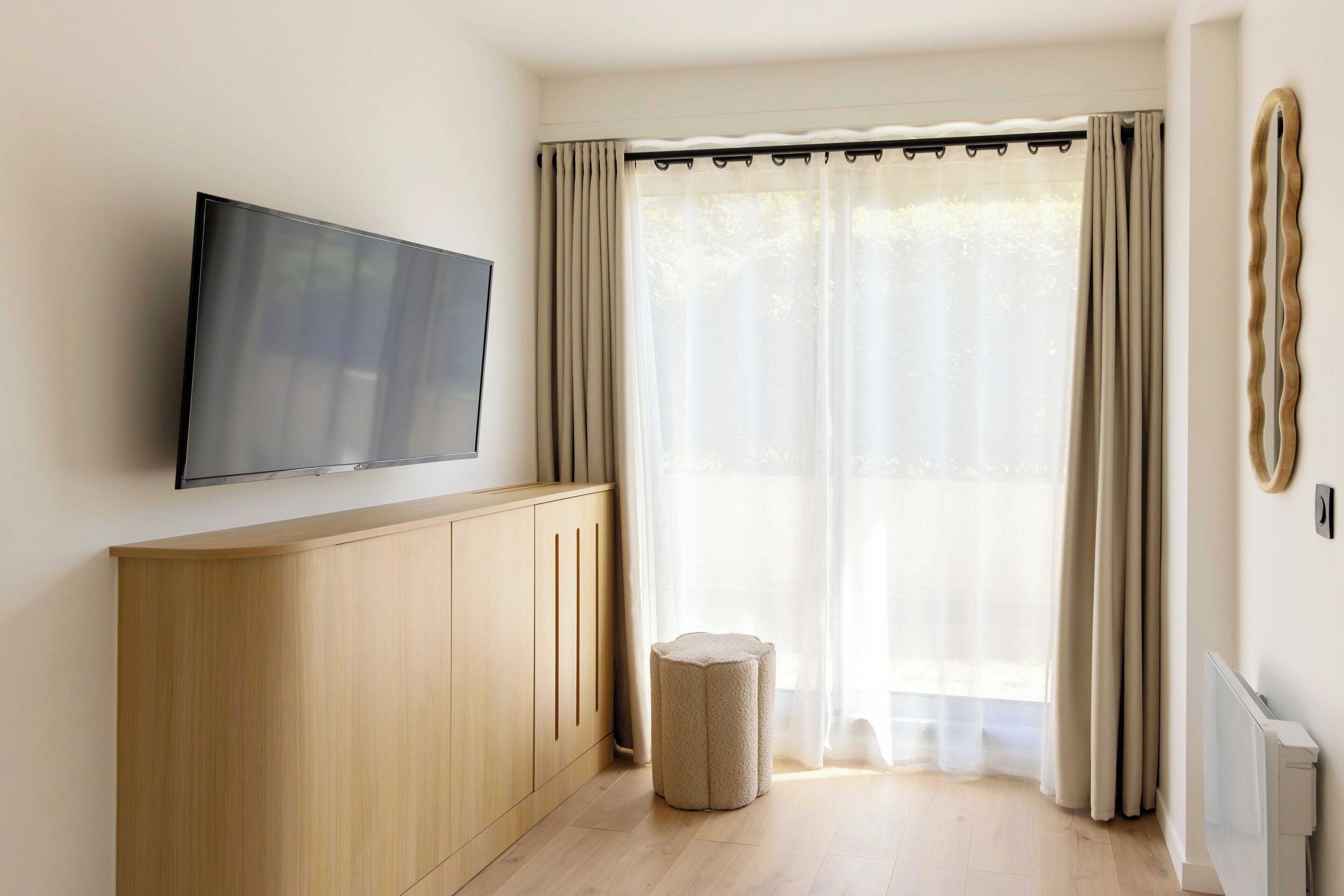 Una habitación con una televisión de pantalla plana montada en la pared, un mueble de madera, un otoman redondo y un ventanal con cortinas ligeras.