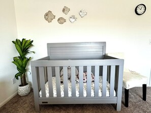 A gray crib with a patterned mattress, a potted plant, and wall decorations including a clock. - Spring Valley Villa (Las Vegas)