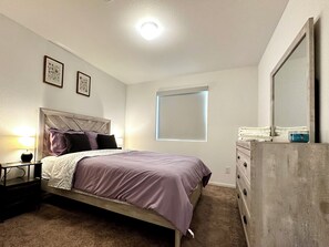 Une chambre à coucher comprenant un lit, une table de chevet, une commode et une fenêtre avec des stores.